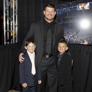 Michael Bisping with family celebrating at the UFC Hall of Fame ceremony. Bright stage lights, happy faces, backdrop filled with UFC logos and imagery.