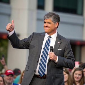 Sean Hannity, standing confidently in front of a crowd. Wearing a gray suit, white shirt, and blue-striped tie. Thumbs up gesture.
