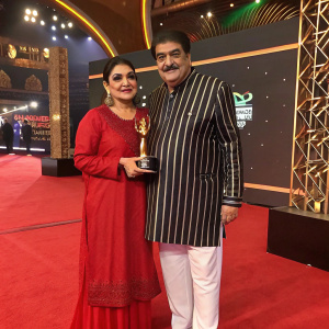 Raza Murad and Samina Murad at a glamorous awards ceremony. Raza in a classic striped kurta paired with white pants. Samina in an elegant traditional red dress. Both smiling for the camera.