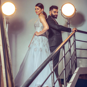 Virat Kohli, in a black suit, stands alongside Anushka Sharma at the Bollywood Filmfare Awards. She wears a stunning white gown with intricate embroidery. They pose for photographers against a backdrop of bright stage lights.