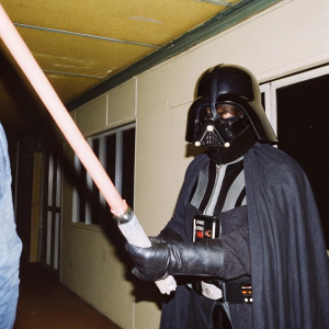 Darth Vader in an intimidating pose. His black helmet with red glowing eyes stands out against a dimly lit, ominous background.