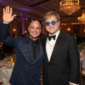 Andre Agassi and Elton John at a benefit gala, smiling and waving at the camera. Lavish ballroom setting with chandeliers.