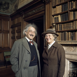 Vintage photograph of Albert Einstein and Elsa Lowenthal Einstein in a cozy library. Both smiling warmly. Albert Einstein stands on the left, with a mustache, while Elsa wears a hat and stands on the right. The background features ornate wooden bookshelves and a fireplace.
