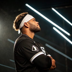 Yoan Moncada, Chicago White Sox player, stands with a rugged look. Black jersey, white headband. Confident posture. Dark backdrop with light trails, creating a moody atmosphere