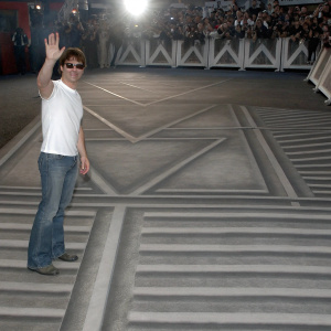 Tom Cruise, in casual attire, at a film premiere. Jeans and white t-shirt. Crowd and flashing cameras in background.