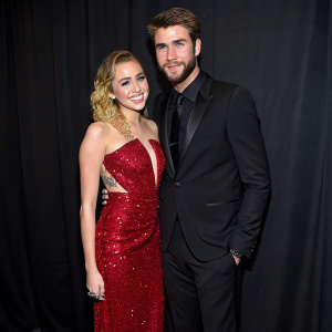 Miley Cyrus in a sparkling red dress, paired with Liam Hemsworth in a traditional black suit. The couple stands against a plain black background at a gala event.