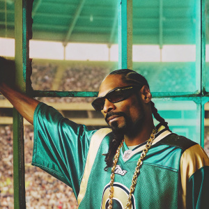 Snoop Dogg, sporting a full beard and cornrows, poses confidently. He is dressed in a LA Rams jersey and gold chains. The background is a blurred out audience at a stadium.