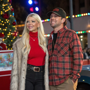 Tara Reid and Ted Dhanik at the 86th Annual Hollywood Christmas Parade. Tara wearing a red turtleneck top, fur stole, and belt. Ted in a plaid shirt and cap. Festive backdrop.