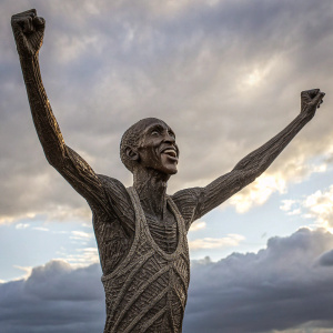 Three-dimensional woven sculpture of Mo Farah. Celebrating victory with arms raised. Dark material, textured weave, cloudy sky backdrop.