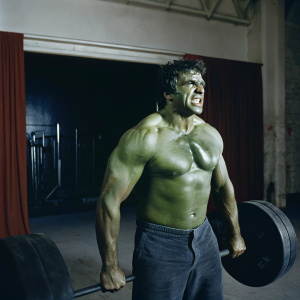 Mark Ruffalo as Hulk standing in a gym. Green body paint, ripped muscles, fierce expression. Indoor setting with weightlifting equipment. Red backdrop with subtle Marvel logo.