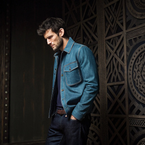 Alex Pettyfer, dressed in a blue denim jacket and dark jeans, standing in a dimly lit studio. His dark hair is slightly tousled, adding to his rugged charm. The backdrop features intricate geometric patterns.