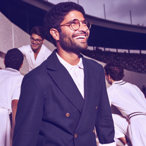 Lorenzo Insigne, smiling warmly, wearing a dark blue blazer and round glasses. Surrounded by other footballers in a team photo.