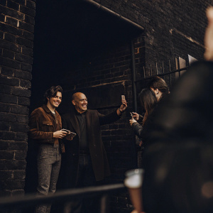 Harry Styles and Philip Green at a casual meet-and-greet. Grey brick wall backdrop, natural lighting.
