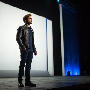 Zac Efron, standing alone on a dimly lit stage, the background a stark white with occasional flickers of blue. A dramatic cinematic scene, showcasing his role as the Human Torch.