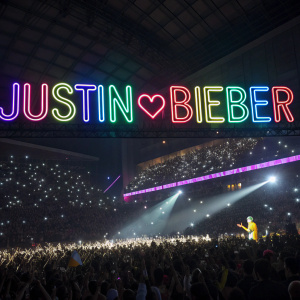 Large neon sign displaying Justin Bieber's name. Crowded stadium, fans waving light sticks