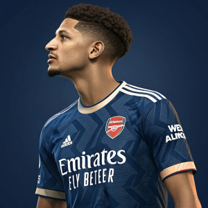 William Saliba, Arsenal soccer player, wearing a navy blue jersey. The jersey has white sponsor logos and team branding. He has short, curly hair. Dark blue background, studio setting
