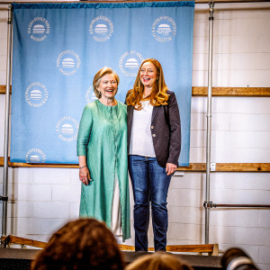 Hillary Clinton and Chelsea Clinton at a fundraising event. Hillary in green dress, Chelsea in white shirt and dark blazer with jeans. Against a blue backdrop with Clinton Foundation logos.