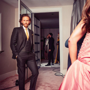 Tom Hiddleston in a sleek black suit with a vibrant yellow tie, standing in a bustling backstage area at a high-profile awards show.