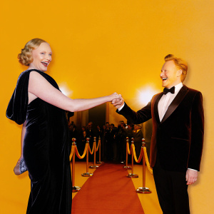 Gwendoline Christie and Conan O'Brien smiling and shaking hands at award ceremony. Red carpet event with flashing cameras.