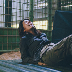 Grace Park, portraying Lieutenant Sharon 'Boomer' Valerii, in a dramatic scene from Battlestar Galactica. Lying on a metal surface, looking distressed. Background features large cage structures. Dark lighting enhances the somber mood.