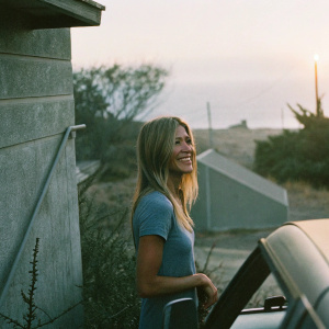 Jennifer Aniston, blonde actress, in a casual blue top, standing outdoors against a sunset backdrop. Smiling for a candid photo shoot.