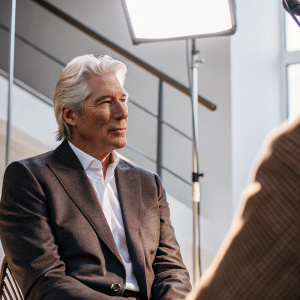 Richard Gere in a formal interview setting. Wears a dark grey suit and white shirt. Bright studio lights illuminate his face and outfit. Background features a sleek, modern design.