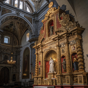 An ornate religious sculpture from the Church of Santa Maria Tonantzintla in Puebla, Mexico. The decoration is rich with intricate gold and silver detailing, red and brown accents, and layers of figures and designs creating a complex visual effect.
