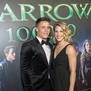 Colton Haynes and Emily Bett Rickards at a gala event celebrating the 100th episode of 'Arrow'. The duo poses in front of a dazzling backdrop featuring the show's logo and cast pictures.