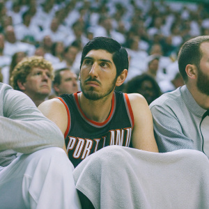 Enes Kanter, in Portland Trail Blazers jersey, sitting on the bench during a NBA game. Serious expression. The crowd is visible in the background.