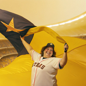 Sarah, fan of the Houston Astros, posing in a stadium. Holding a big Astros flag. Crowd in the background