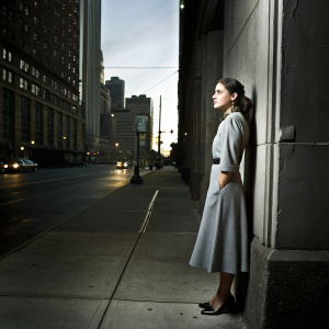 Sofia Black D'Elia, standing alone on a city street at dusk, wearing a long gray dress and black heels.