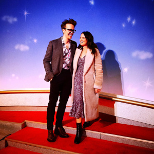 Tom Fletcher and Giovanna Falcone at the Cirque du Soleil premiere. Tom in a dark grey blazer, patterned shirt, and black slacks. Giovanna in a taupe coat with fur trim, paired with a patterned dress. They both share a warm smile against a sleek blue backdrop adorned with white accents.