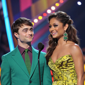 Daniel Radcliffe and Vanessa Minnillo at a vibrant award ceremony. Daniel in a stylish green suit, Vanessa adorned with large earrings. The stage is filled with colorful decorations and vibrant lighting.