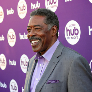 Ernie Hudson, wearing a grey suit and lavender shirt, smiling at the camera on the Hub TV Network sponsored event. Repetitive Hub TV Network logos on a purple backdrop.