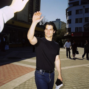 Henry Cavill, in a casual black t-shirt and jeans, waving to fans in a bustling city square.