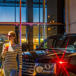 Emma Roberts enjoying a casual day at a luxury car showroom. She's wearing a trendy striped shirt and sunglasses, holding a drink. A shiny Land Rover is parked nearby under the sunny sky.