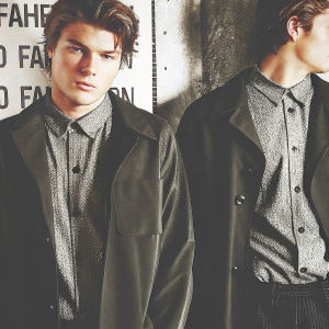 Jordan Barrett, a young man with neatly styled hair, is photographed in a studio setting. He is dressed in a black jacket over a textured shirt. The background is stark white with black text elements. This is a close-up shot focusing on his upper torso and face. The lighting is even, highlighting the textures of his clothing and facial contours. The image is modern and straightforward.
