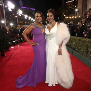 Vivica A. Fox and Rashel Pouri at a glitzy award show. Both in elegant gowns. Vivica in purple, Rashel in white with fur accents. Crowds and photographers surround them on the red carpet.