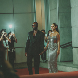 Azuka Ononye and Alesha Dixon at a high-profile awards show. Azuka in a sleek black suit, Alesha in a silver sequined dress. Flashing cameras and fans in the background.