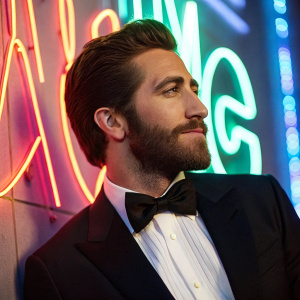 Jake Gyllenhaal, wearing a bow tie and a beard, posing against a vibrant neon background.