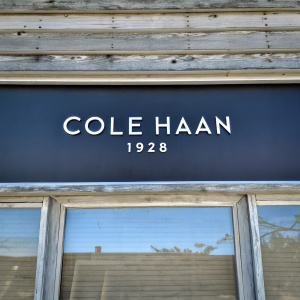 Cole Haan storefront sign. Vintage look, dark navy backdrop, white text 'Cole Haan 1928', wooden texture facade
