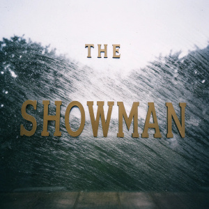 Gilded lettering of 'The Greatest Showman'. Textured gold font against crisp white backdrop, elegant and stylish design with subtle brushstroke effect.