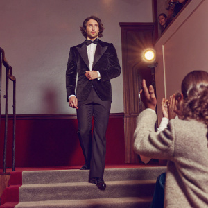 Aaron Taylor-Johnson at the premiere. Wearing a black tuxedo and bow tie, standing confidently on a brightly lit stage with fans cheering in the background.