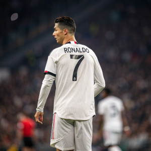 Photograph of Cristiano Ronaldo. Back view. White jersey with 'RONALDO 7' visible. Blurred crowd at Manchester United stadium.