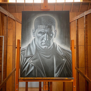Realistic drawing of Frank Castle, aka the Punisher, by an artist. The drawing is in grayscale and detailed. The artist is showing the artwork in a studio with a plain background.