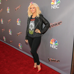 Christina Aguilera at the launch event for The Voice season 8. Wearing a casual black jacket over a graphic t-shirt and black leggings, red heels. Background filled with event branding.