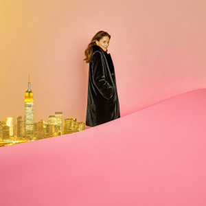 Emily DiDonato, elegant pose, in a black velvet coat, against the backdrop of a vibrant city skyline at night.