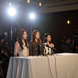 Areum, Hyomin, and Jiyeon from T-ara N4. They're at a press conference. White tableclothed table behind them. Bright lights and microphones in the foreground.