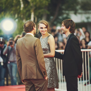 Cara Delevingne, Jake Schreier, and Nat Wolff at the film festival premiere. Standing on a red carpet. Silver sequined dress, brown suit, black suit respectively. Crowd of photographers and fans in the background.