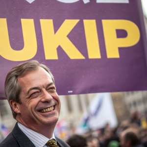Close-up shot of UKIP campaign poster. Nigel Farage smiling. Purple background with yellow text. Blurry crowd in background.